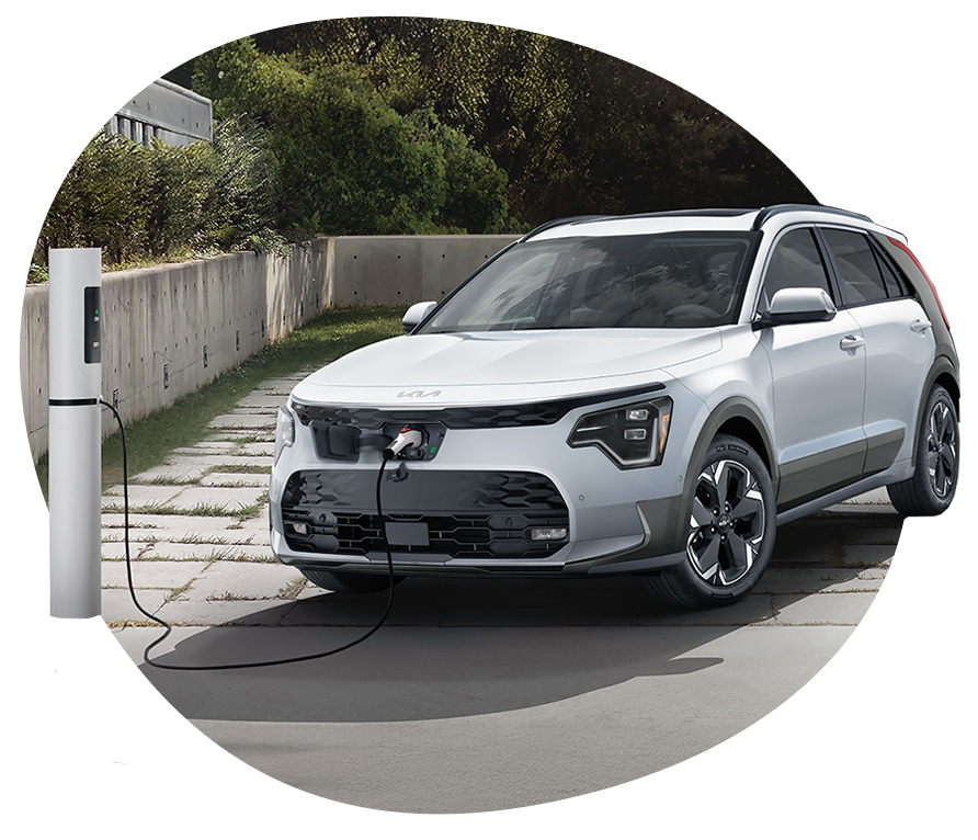 Kia Niro EV Charging Outdoors White Kia Niro EV plugged into a sleek outdoor charging station on a paved stone path, with a concrete wall and greenery in the background.